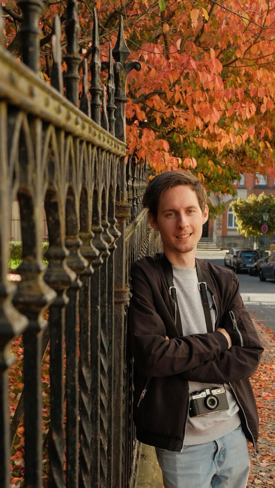Portrait of Michele leaning on an iron fence in autumn, with a film camera around his neck